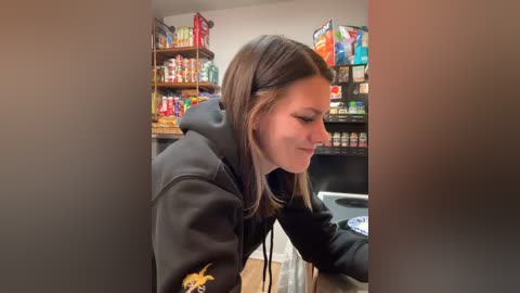 Media: A video of a young woman with straight brown hair, wearing a black hoodie, sitting at a gaming setup with colorful shelves filled with snacks and gaming items in the background.