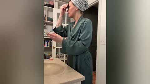 Media: A video of a woman with a light complexion and light brown hair in a gray checkered turban, wearing a green bathrobe, applying makeup in a bathroom with beige countertops, white cabinets, and toiletries on shelves.