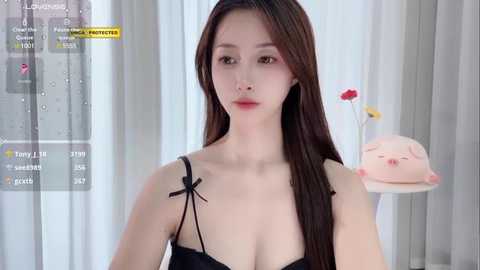 Media: A video of an East Asian woman with long, straight brown hair, wearing a black strapless top, standing in a modern, minimalistic room with white curtains and a pink pillow.