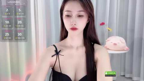 Media: Video of an East Asian woman with fair skin, long black hair, and red lipstick, wearing a black bra with a bow. She stands in a minimalist room with a white wall and a stuffed animal.