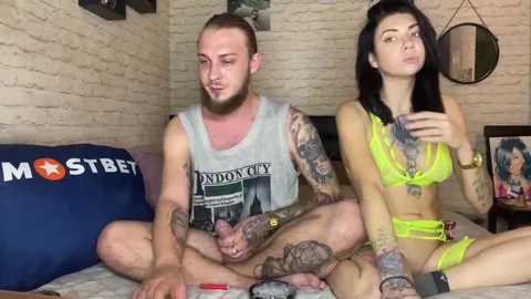 Media: Video of a tattooed man in a gray tank top and a woman in neon yellow lingerie, sitting on a bed with brick walls and London-themed decor.