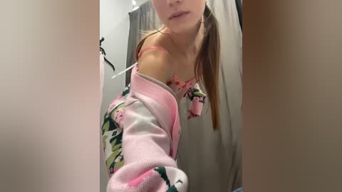 Media: A video of a light-skinned woman with long brown hair in a ponytail, wearing a pink and white floral robe, standing in a bathroom with beige walls and a shower curtain.