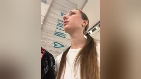 Media: Video of a young woman with long brown hair tied in pigtails, wearing a white shirt and silver earrings, standing in a brightly lit room with a white ceiling fan and a \"Be Positive\" sign.