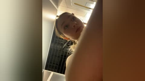 Media: A video of a young, fair-skinned woman with blonde hair, looking up through a narrow gap. She has a neutral expression. Background shows a modern, dimly lit room with metal bars.