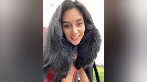 Media: Video of a smiling woman with long black hair, wearing a black fur-trimmed coat, in a blurred outdoor setting.