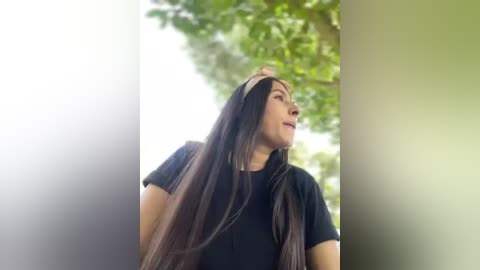 Media: Video of a young woman with long, straight, dark brown hair, wearing a black shirt, standing outdoors against a blurred, green, leafy background, looking up thoughtfully.