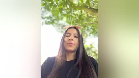 Media: Video of a young woman with long, straight dark brown hair, wearing a black shirt, standing outdoors against a blurred green background of trees.