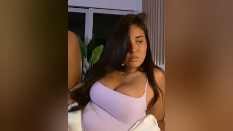 Media: Video of a curvy, Latina woman with long, dark hair, wearing a low-cut, light purple top, sitting in a dimly lit room with a potted plant and window visible in the background.