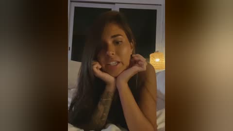 Media: Video of a young woman with long brown hair, light skin, and a small tattoo on her left arm, resting her chin on her hands in a dimly lit bedroom.