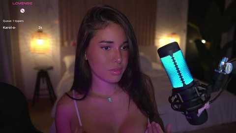 Media: Video of a young Asian woman with long black hair, wearing a pink camisole, in a dimly lit room with a green screen and a bright blue microphone.