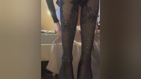 Media: Video of a woman's legs in black fishnet stockings with sequins, wearing a black lace top, standing in a dimly lit bedroom.