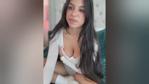 Media: Video of a young woman with long black hair, light skin, and full lips, wearing a white cardigan that reveals a white bra. She is sitting on a green bed with a tufted headboard, indoors.