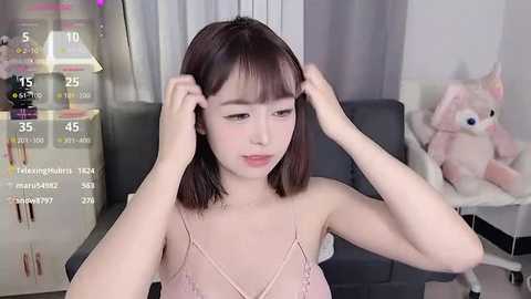 Media: Video of a fair-skinned East Asian woman with straight, shoulder-length brown hair, adjusting her bangs in a modern living room with a plush pink cat toy and digital game stats displayed.