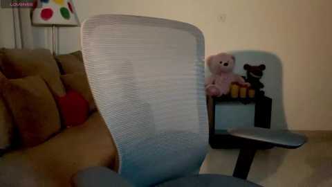 Media: Video of a beige sofa with a stuffed bear and a blue mesh office chair, set against a cream-colored wall with a colorful lamp and a black shelf with a wooden toy in the background.