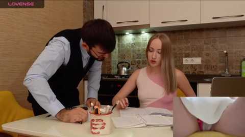 Media: Video of a young Caucasian woman in a pink dress and a man in a vest and glasses, cutting a cake in a modern kitchen.