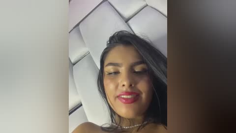 Media: Video of a young Latina woman with long black hair, wearing red lipstick, against a white, quilted headboard. She has a light brown complexion and is smiling.