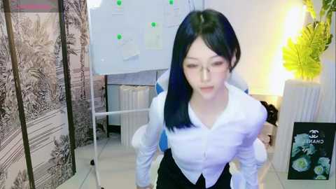 Media: A video of an East Asian woman with long black hair, wearing a white blouse and black skirt, standing in a modern office with a whiteboard, plant, and decorative screens.