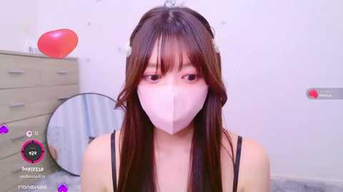 Media: Video of an Asian woman with long brown hair, wearing a pink mask and black tank top, sitting on a gray floor in a minimalist room with a white wall, a light-colored dresser, and a round mirror.