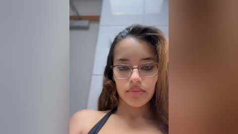 Media: A video of a young Latina woman with long, wavy brown hair, wearing glasses, black top, and light skin, standing in a bathroom with white tiles and a wooden shelf.