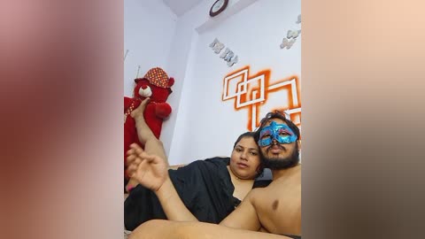 Media: Video: A middle-aged Indian woman with medium skin tone and a man with a beard and blue mask, both topless, sitting on a couch. Red clay figurine and orange wall art in background.