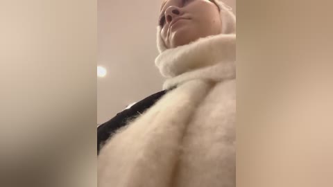 Media: A low-angle video of a woman in a fluffy white coat, with a hint of a black garment underneath. She has a calm expression, and the background is blurred and beige, possibly indoors.