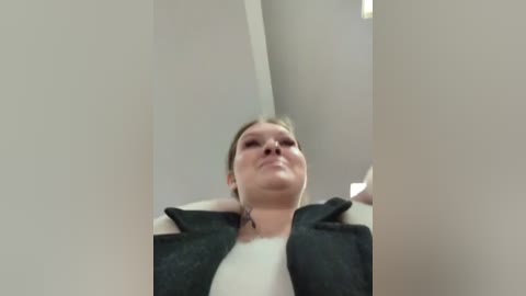 Media: A video of a smiling, light-skinned woman with short brown hair, wearing a black jacket over a white shirt, captured from a low angle, partially obscured by vertical white walls.