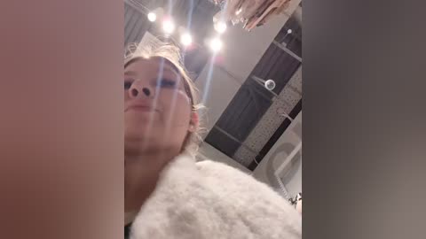 Media: Video of a blonde woman in a fluffy white bathrobe, standing in a bathroom with a tiled wall and mirror. The image has a soft focus and a warm, cozy ambiance.