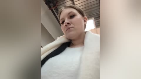 Media: Video of a young woman with light skin and brown hair, wearing a white and black fleece jacket, standing indoors under a corrugated metal roof.