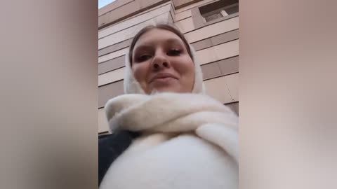 Media: Video of a young woman with fair skin and light brown hair, wearing a white fluffy scarf and a black jacket, standing against a beige brick building. She has a neutral expression.