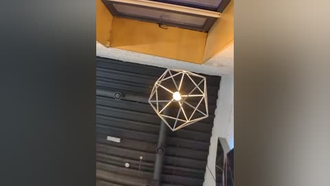 Media: Video of a modern, minimalist room with a large, geometric, metal pendant light hanging from a dark, ribbed ceiling. The walls are painted in warm tones of yellow and beige.