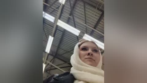 Media: Video of a young Caucasian woman with fair skin and light brown hair, wearing a white hijab and a dark jacket, standing in an industrial warehouse with a corrugated metal ceiling and large, rectangular windows.