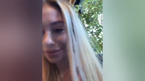 Media: Video of a smiling young woman with long, straight blonde hair, fair skin, and a slight tan. She's wearing a sleeveless top. Background shows a blurry green leafy tree.