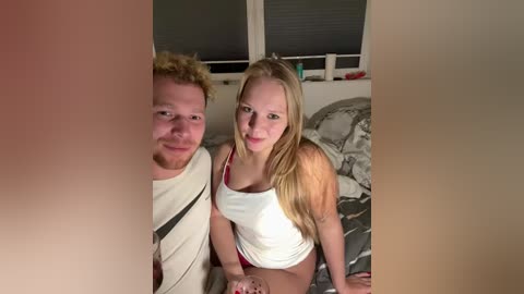 Media: Video of a blonde woman in a white tank top and red shorts, sitting next to a man with curly hair in a striped shirt, on a bed with grey bedding.