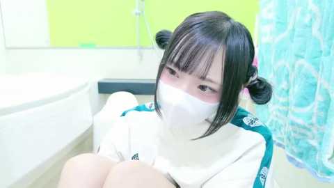 Media: Video of an Asian woman with black hair styled in buns, wearing a white face mask, sitting on a bathtub ledge in a brightly lit bathroom with a green wall and turquoise shower curtain.