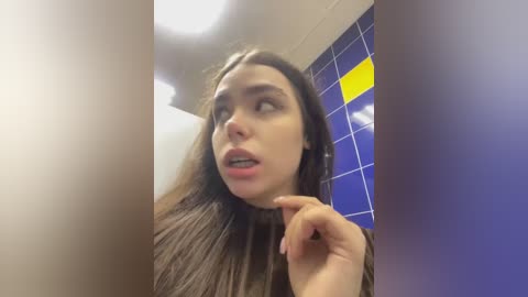 Media: Video of a young woman with long brown hair, light skin, and expressive eyes in a tiled bathroom, with a surprised expression.