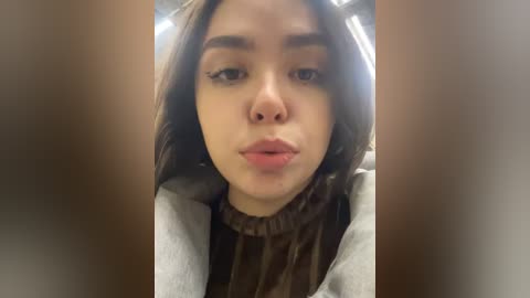 Media: Video of a young woman with medium brown skin, long dark hair, and full lips, puckered for a kiss, wearing a gray coat. Background blurred, indoors, casual setting.