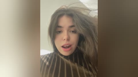 Media: Video of a young woman with long, wavy brown hair, wearing a striped top, captured in a casual, candid moment. She has a neutral expression, with slightly open lips, and is partially obscured by a vertical, out-of-focus object.