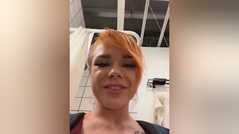 Media: Video of a fair-skinned woman with short, dyed orange hair, wearing a black and purple top, smiling, standing in a bathroom with white tiles, a towel rack, and a window.