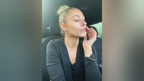 Media: Video of a blonde woman with her hair tied up, wearing a black cardigan, eating a piece of food with her eyes closed, seated in a car.