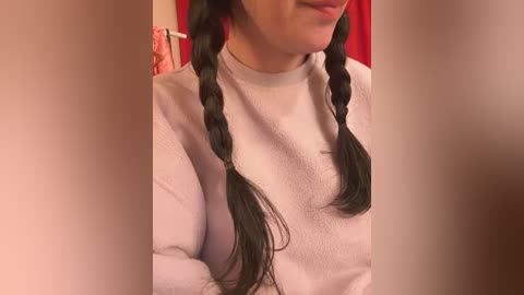 Media: Video of a woman with fair skin, wearing a white long-sleeve shirt, featuring two braided black ponytails. Background features a red curtain and a pink towel, suggesting a cozy, indoor setting.