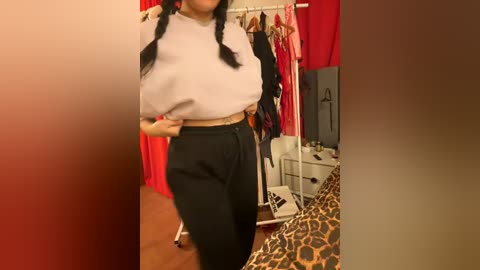 Media: Video of a woman with long black hair styled in braids, wearing a white cropped top and high-waisted black pants, standing in a room with a leopard-print bedspread, red curtains, and a clothing rack.