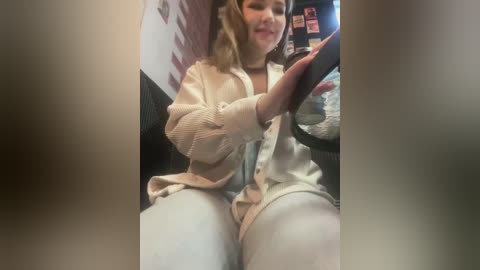 Media: Video of a smiling, light-skinned woman with shoulder-length blonde hair, wearing a beige sweater and pants, sitting in a car, holding a phone.