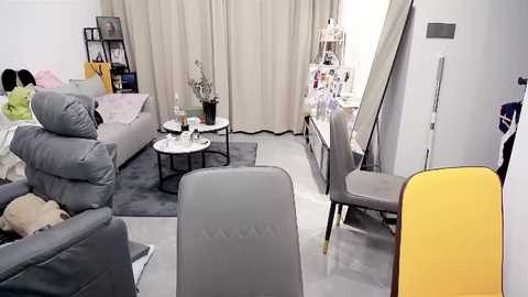 Media: Video of a modern, minimalist living room with beige curtains, grey sofa, yellow chair, wooden coffee table, and grey carpet.