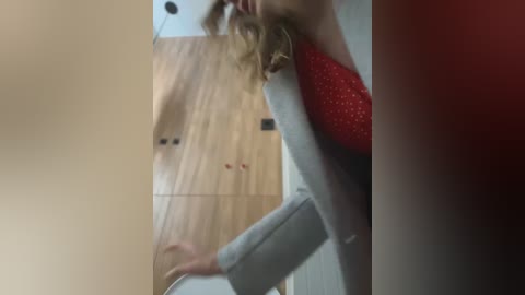 Media: Video of a blonde woman with wavy hair, wearing a red top and grey pants, bending over to adjust a white chair, in a modern room with light wood walls and a red accent.