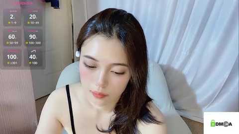 Media: Video of a young East Asian woman with long, straight brown hair, wearing a black spaghetti strap top, lying on a white bed, with a digital health app overlay showing 20\u00b0C, 60% humidity, and 90% humidity.