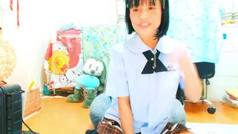 Media: Video of a cheerful Asian woman with straight black hair, wearing a light blue blouse and plaid skirt, seated at a cluttered wooden desk in a colorful, childlike room.