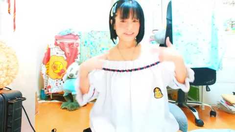 Media: Video of a smiling Asian woman with short black hair, wearing a white off-shoulder blouse with colorful embroidery, standing in a cluttered room with a blue chair, a wicker basket, and a desk.