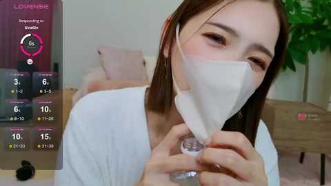 Media: Video of a young Asian woman with long brown hair, wearing a white mask, holding a glass, in a cozy, modern bedroom with a wooden nightstand and green plant.