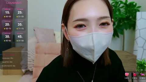 Media: Video of an East Asian woman in a black turtleneck, wearing a white face mask, with brown hair, indoors, displaying a \"Love Live!\" virtual game screen with health and energy bars.