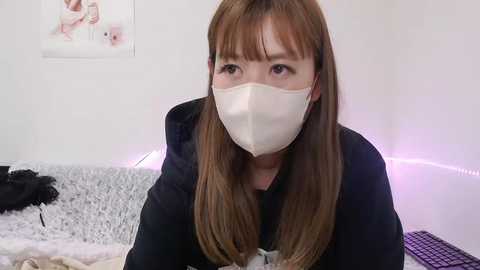 Media: Video of a young Asian woman with long brown hair, wearing a white mask and black hoodie, sitting on a gray plush bed. Background features a white wall with a framed photo and purple LED strip lighting.